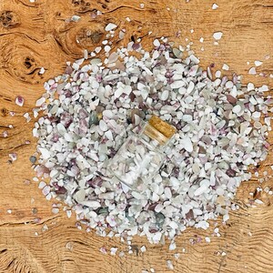 May include: A small glass bottle with a cork stopper filled with white and pink gemstone chips. The bottle is lying on a pile of similar chips on a wooden surface.