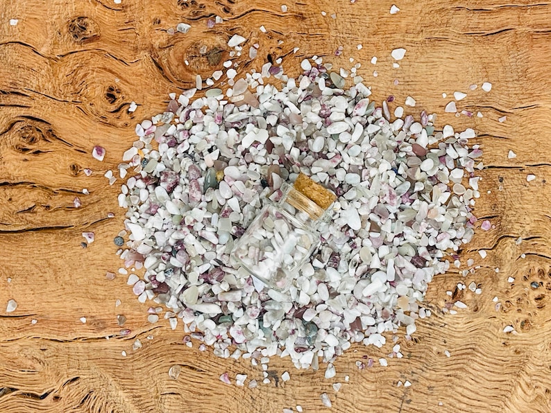 May include: A small glass bottle with a cork stopper filled with white and pink gemstone chips. The bottle is lying on a pile of similar chips on a wooden surface.