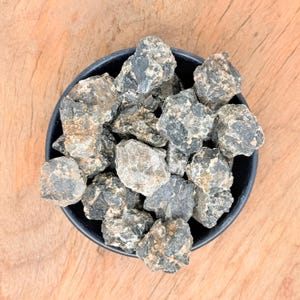 May include: A black bowl filled with rough, dark gray and brown stones. The stones have a textured appearance and varying sizes. The bowl is set against a light brown wooden surface.