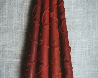 May include: Four red taper candles with rose and vine detailing. The candles are arranged vertically against a neutral linen background. The candles are approximately 10 inches tall.