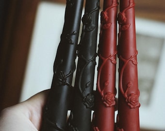 May include: Four taper candles, two black and two red, are decorated with rose and vine designs. The candles are held in a hand, with the wicks visible at the top. The candles are a decorative item.