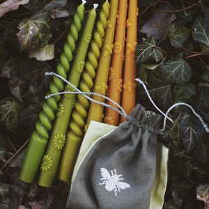 May include: A collection of decorative candles in shades of green, yellow, and orange, with floral and spiral designs. The candles are accompanied by a small gray drawstring bag with a white bee emblem. The background features green leaves.