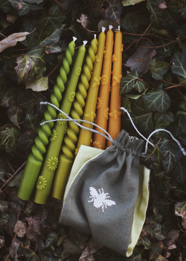 May include: A collection of decorative candles in shades of green, yellow, and orange, with floral and spiral designs. The candles are accompanied by a small gray drawstring bag with a white bee emblem. The background features green leaves.
