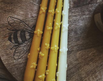 May include: Four beeswax taper candles in shades of yellow and white, decorated with bee-shaped embellishments. The candles are arranged on a wooden tray with a bee illustration. A rustic and natural home decor item.