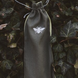 May include: A dark olive green fabric drawstring pouch featuring a white embroidered bee design on the front. The pouch is cinched at the top with a thin, light-colored string and has a light green interior lining visible at the edges.