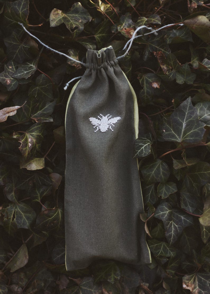 May include: A dark olive green fabric drawstring pouch featuring a white embroidered bee design on the front. The pouch is cinched at the top with a thin, light-colored string and has a light green interior lining visible at the edges.