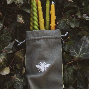 May include: A collection of decorative candles in shades of green and yellow, some with floral and bee designs, are displayed in a gray drawstring bag with a white embroidered bee. The candles are set against a backdrop of green leaves.