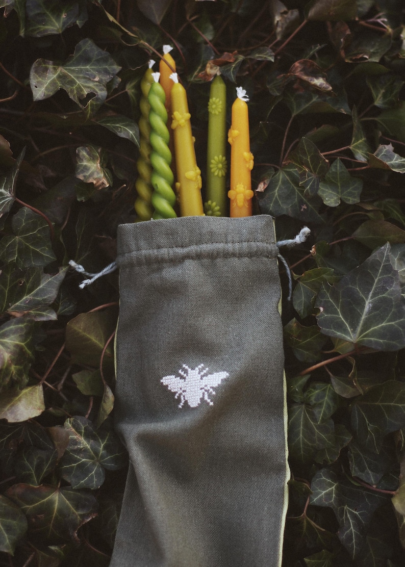 May include: A collection of decorative candles in shades of green and yellow, some with floral and bee designs, are displayed in a gray drawstring bag with a white embroidered bee. The candles are set against a backdrop of green leaves.