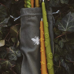 May include: A dark gray fabric drawstring bag featuring a white embroidered bee icon, containing several taper candles. The candles include green and orange colors with embossed flower and bee details, and one green spiral-shaped taper candle.