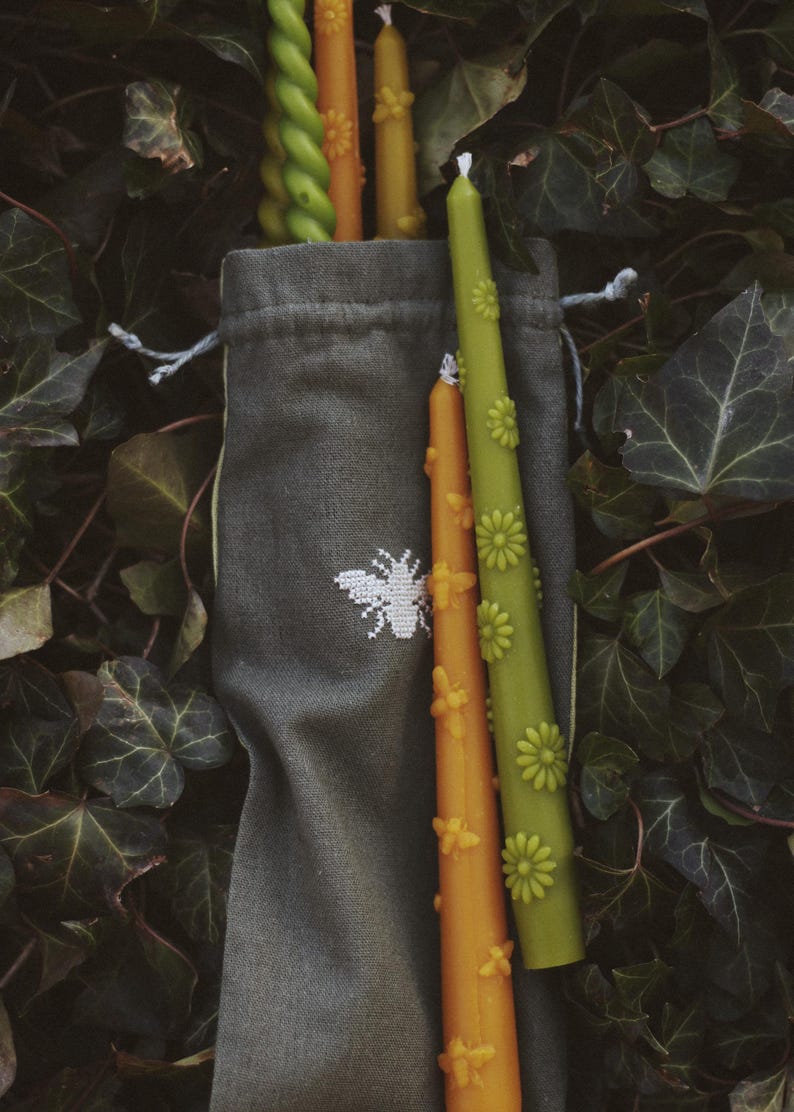 May include: A dark gray fabric drawstring bag featuring a white embroidered bee icon, containing several taper candles. The candles include green and orange colors with embossed flower and bee details, and one green spiral-shaped taper candle.