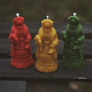 Puede incluir: Tres velas decorativas con forma de figuras, cada una con una mecha. Las velas son de color rojo, amarillo y verde. Las figuras visten ropa detallada y sostienen pequeños objetos. Las velas están colocadas sobre una superficie de madera oscura.