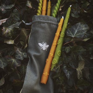 May include: A collection of decorative candles in various colors, including orange, green, and yellow, are displayed. Some candles feature floral and bee designs. The candles are presented in a gray drawstring bag with a white bee embroidery, set against a backdrop of green foliage.