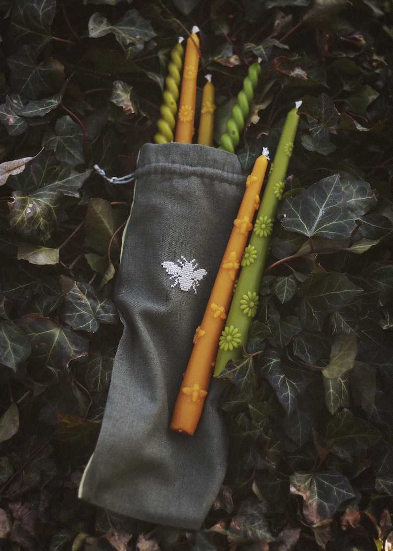 May include: A collection of decorative candles in various colors, including orange, green, and yellow, are displayed. Some candles feature floral and bee designs. The candles are presented in a gray drawstring bag with a white bee embroidery, set against a backdrop of green foliage.