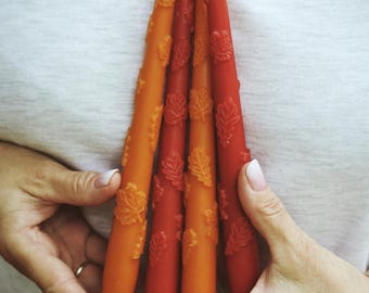 May include: A set of four tapered candles in shades of orange and red, each featuring a textured, raised relief pattern of small oak leaves along the surface. The candles are arranged vertically and held together in a cluster.