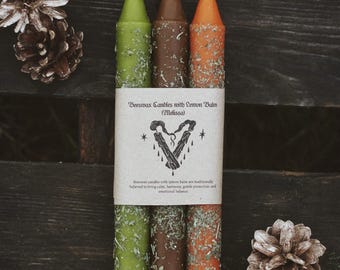 May include: A set of three beeswax candles with lemon balm in green, brown, and orange, adorned with dried herbs. The candles are wrapped with a label that reads "Beeswax Candles with Lemon Balm (Melissa)". The candles are displayed on a dark wooden surface.