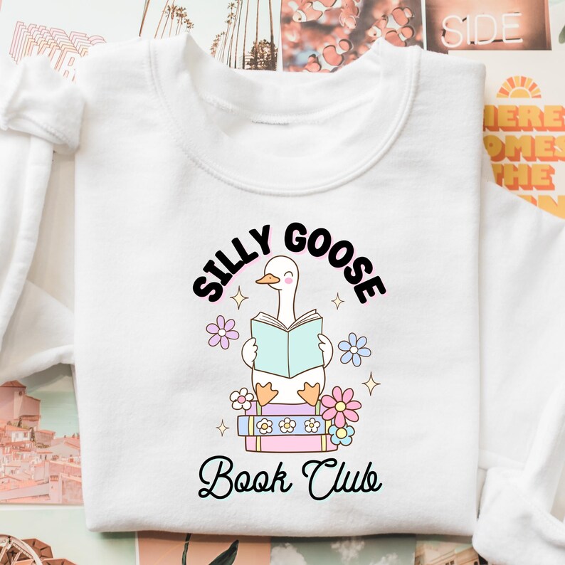 K&ouml;nnte beinhalten: Wei&szlig;es Sweatshirt mit einem Grafikdruck einer Comic-Gans, die ein blaues Buch tr&auml;gt und auf einem Stapel B&uuml;cher sitzt. Der Text "Silly Goose Book Club" ist unter der Gans aufgedruckt.