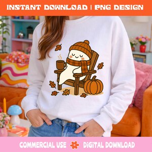 Pode incluir: Camisola branca com um fantasma de desenho animado usando um chap&eacute;u e cachecol marrons, sentado em uma cadeira de madeira, segurando uma caneca e cercado por folhas de outono e uma ab&oacute;bora. O texto na parte superior diz "INSTANT DOWNLOAD | PNG DESIGN".