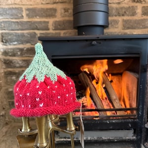 May include: A hand-knitted hat shaped like a strawberry, featuring a bright red body with small white stitched details and a light green pointed top. The hat has a rolled brim and is displayed on a brass stand with a small attached paper tag.