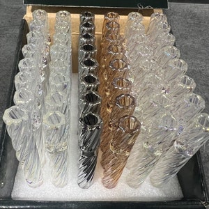 May include: A display of glass smoking pipes in a variety of colors, including clear, silver, and gold. Each pipe has a spiral design and a flared mouthpiece. The pipes are arranged in rows inside a black box.