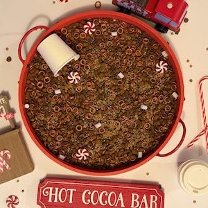 May include: A Christmas-themed arrangement with a red tray filled with cocoa ingredients. Includes a wooden hot chocolate stand, a red truck, peppermints, candy canes, and a "Hot Cocoa Bar" sign.