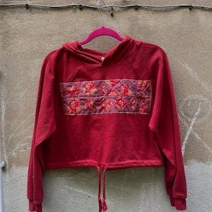 Puede incluir: Sudadera roja de capucha corta con un diseño de patchwork en la parte delantera. La sudadera tiene un cordón en la parte inferior y una capucha.