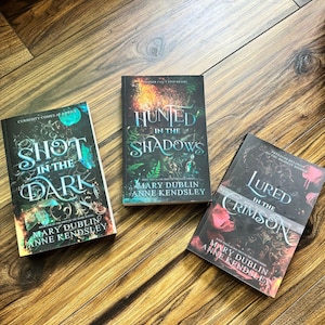 Pode incluir: Três livros de capa dura com capas escuras são exibidos em uma superfície de madeira. Os títulos são "Shot in the Dark", "Hunted in the Shadows" e "Lured in the Crimson". Os autores são Mary Dublin e Anne Kendsley.