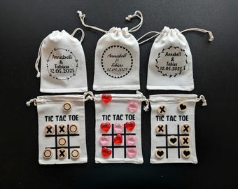 May include: Six off-white fabric drawstring pouches. Three pouches feature a circular wreath design with the text Annabell & Tobias 12.05.2025. Three pouches feature a printed tic tac toe grid with wooden tokens marked with X, O, or heart symbols.