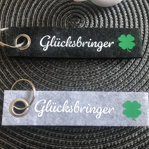 Könnte beinhalten: Zwei schwarze und weiße Filz-Schlüsselanhänger mit einem silbernen Schlüsselring. Beide Schlüsselanhänger haben ein grünes vierblättriges Kleeblatt und den Text "Glücksbringer" in weiß.