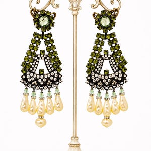 May include: A pair of ornate chandelier earrings featuring dark metal settings, olive green crystals, clear rhinestones, and dangling cream-colored teardrop pearls. The earrings are displayed on a decorative cream-colored metal stand.