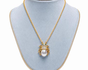 Peut inclure: Un collier doré avec un pendentif en forme de scarabée. Le corps de l'insecte est composé d'une grosse perle blanche, d'un thorax incrusté de cristaux transparents et de deux petites pierres vertes pour les yeux, suspendu à une fine chaîne dorée.