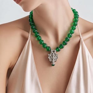 May include: A necklace featuring a strand of round, ribbed, translucent emerald green beads. The necklace includes a central silver-toned metal pendant with an intricate, symmetrical embossed design and a small green bead at the bottom point.
