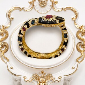 May include: A gold-tone hinged snake bracelet featuring black enamel, clear rhinestones, and inset red and blue faceted stones. The bracelet is displayed on a white and gold decorative stand with ornate floral details and small snake motifs.