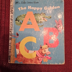 May include: A vintage children's book titled "The Happy Golden ABC" with a blue cover and illustrations of a crocodile, a butterfly, and a cow. The book is part of the "Little Golden Book" series.