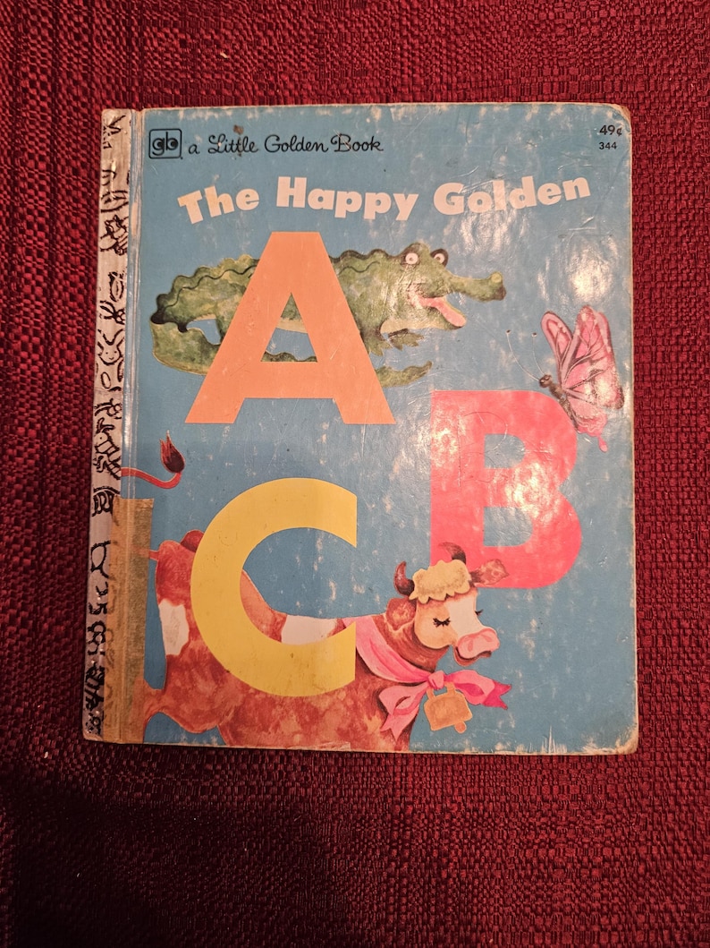 May include: A vintage children's book titled "The Happy Golden ABC" with a blue cover and illustrations of a crocodile, a butterfly, and a cow. The book is part of the "Little Golden Book" series.
