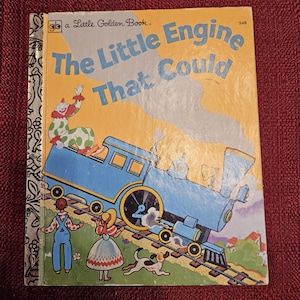 Könnte beinhalten: Ein Vintage Little Golden Book mit dem Titel "The Little Engine That Could" mit einem blauen Zug auf dem Cover. Das Buch hat einen gelben Einband mit einem goldenen Rücken und einem roten Hintergrund.