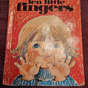 Puede incluir: Una portada de libro infantil vintage con fondo rojo y el título "ten little fingers" en letras negras y doradas. La portada presenta una ilustración de dibujos animados de un niño con cabello rubio y ojos azules, vistiendo una camisa a rayas azules, con ambas manos levantadas en el aire.