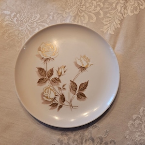 Op de afbeelding: Een wit keramiek bord met een gouden bloemenontwerp met witte rozen en bruine bladeren.