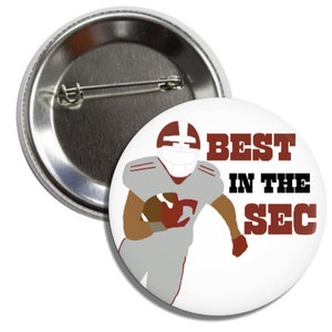 May include: A white button with a cartoon football player wearing a red and gray uniform. The text "BEST IN THE SEC" is printed in black on the button.