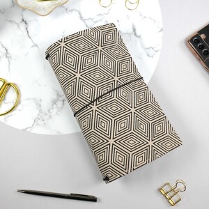 Könnte beinhalten: Ein beiges und schwarzes Notizbuch mit geometrischem Muster und schwarzem Gummibandverschluss. Eine goldene Schere, ein Stift und ein Smartphone sind ebenfalls auf einer weißen Oberfläche zu sehen. Eine kleine Sukkulente befindet sich in der oberen linken Ecke.