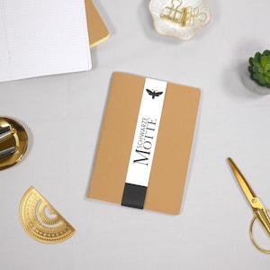 Könnte beinhalten: Ein beiges Notizbuch mit dem Text "SCHWARZE MOTTE" auf einem weißen Band, einer schwarzen Schmetterlingsgrafik und einem schwarzen Band. Goldene Schere, eine goldene Schale mit Stiften und eine kleine Sukkulente sind ebenfalls zu sehen.
