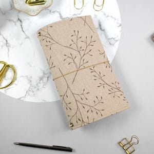 Könnte beinhalten: Ein beiges Notizbuch mit braunem Blumenmuster und einem beigen Gummibandverschluss. Das Notizbuch liegt auf einer weiß-grauen Marmoroberfläche, umgeben von einer goldenen Schere, einem Stift und einem Smartphone.