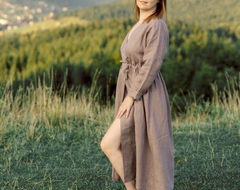May include: A woman in a long, light brown linen dress stands in a field with a mountain range in the background. She is looking at the camera and smiling.
