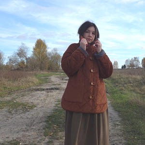 May include: A quilted, rust-colored jacket with a rounded collar and toggle closures. The jacket has a diamond pattern and two front pockets. The person is wearing a long, olive-green skirt. The background shows a dirt path and a cloudy sky.