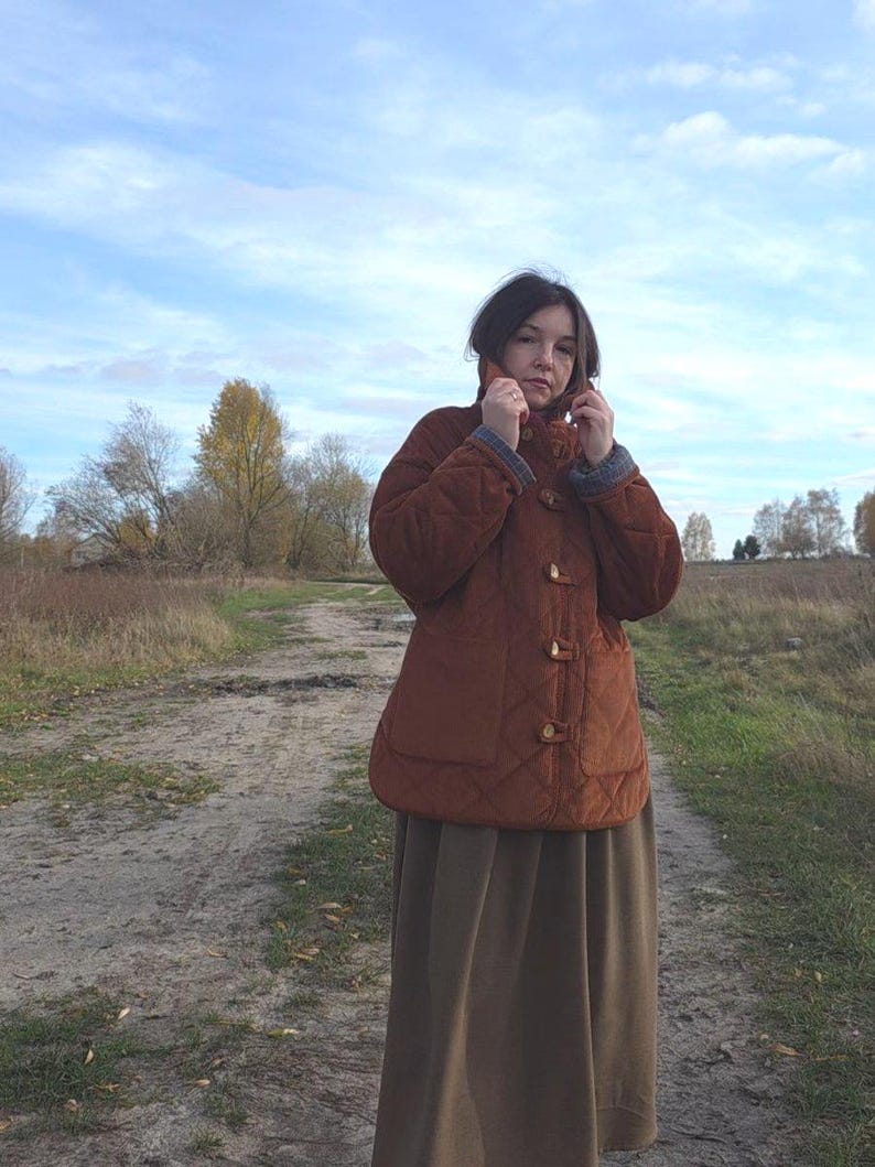 May include: A quilted, rust-colored jacket with a rounded collar and toggle closures. The jacket has a diamond pattern and two front pockets. The person is wearing a long, olive-green skirt. The background shows a dirt path and a cloudy sky.