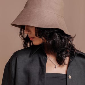 May include: A solid taupe bucket hat with a wide, downward-sloping brim and a structured crown. The hat is crafted from a plain-weave fabric with a visible texture, featuring a horizontal seam detail around the circumference of the crown.