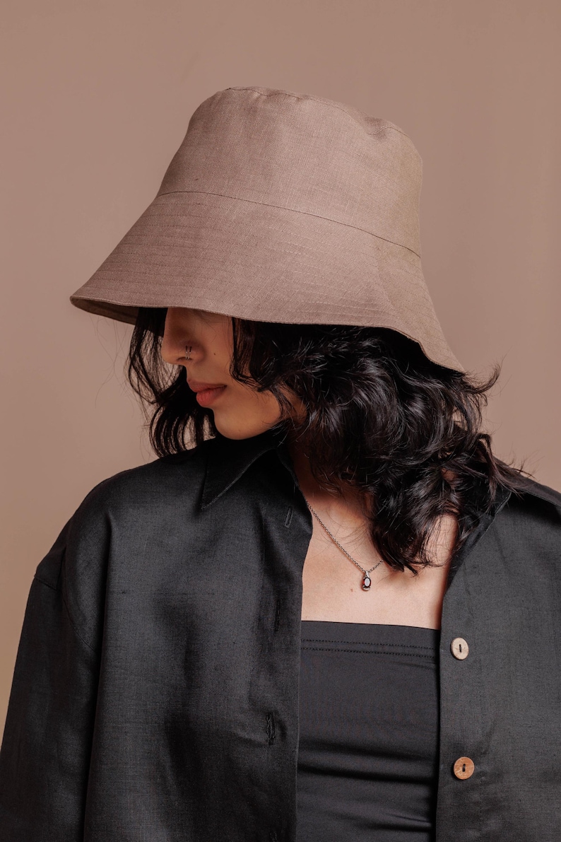 May include: A solid taupe bucket hat with a wide, downward-sloping brim and a structured crown. The hat is crafted from a plain-weave fabric with a visible texture, featuring a horizontal seam detail around the circumference of the crown.