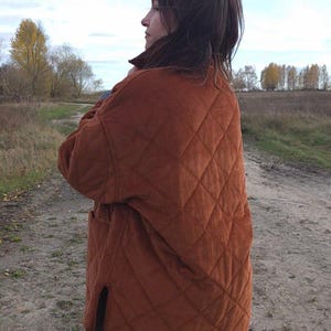 May include: A rust-colored quilted corduroy jacket with a diamond pattern. The jacket has a relaxed fit, a collar, and two front pockets. The person is wearing a brown skirt. The background is a field with trees.
