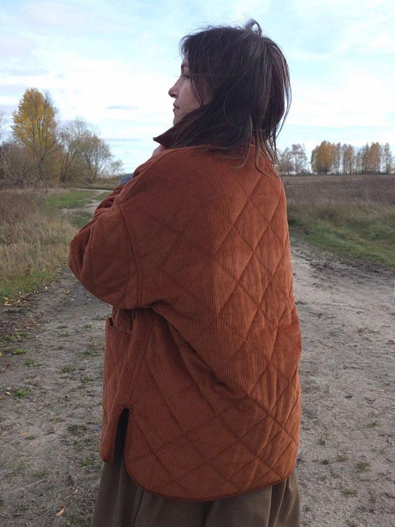 May include: A rust-colored quilted corduroy jacket with a diamond pattern. The jacket has a relaxed fit, a collar, and two front pockets. The person is wearing a brown skirt. The background is a field with trees.