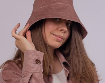 May include: A dusty rose corduroy bucket hat. The hat is paired with a matching corduroy jacket with a buttoned cuff. The jacket is worn over a white ribbed tank top. The background is a solid light gray.