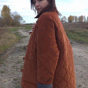 May include: A quilted, rust-colored corduroy jacket with a diamond pattern. The jacket has a button-up front and a plaid lining at the cuffs. The person is standing on a dirt path in a field.